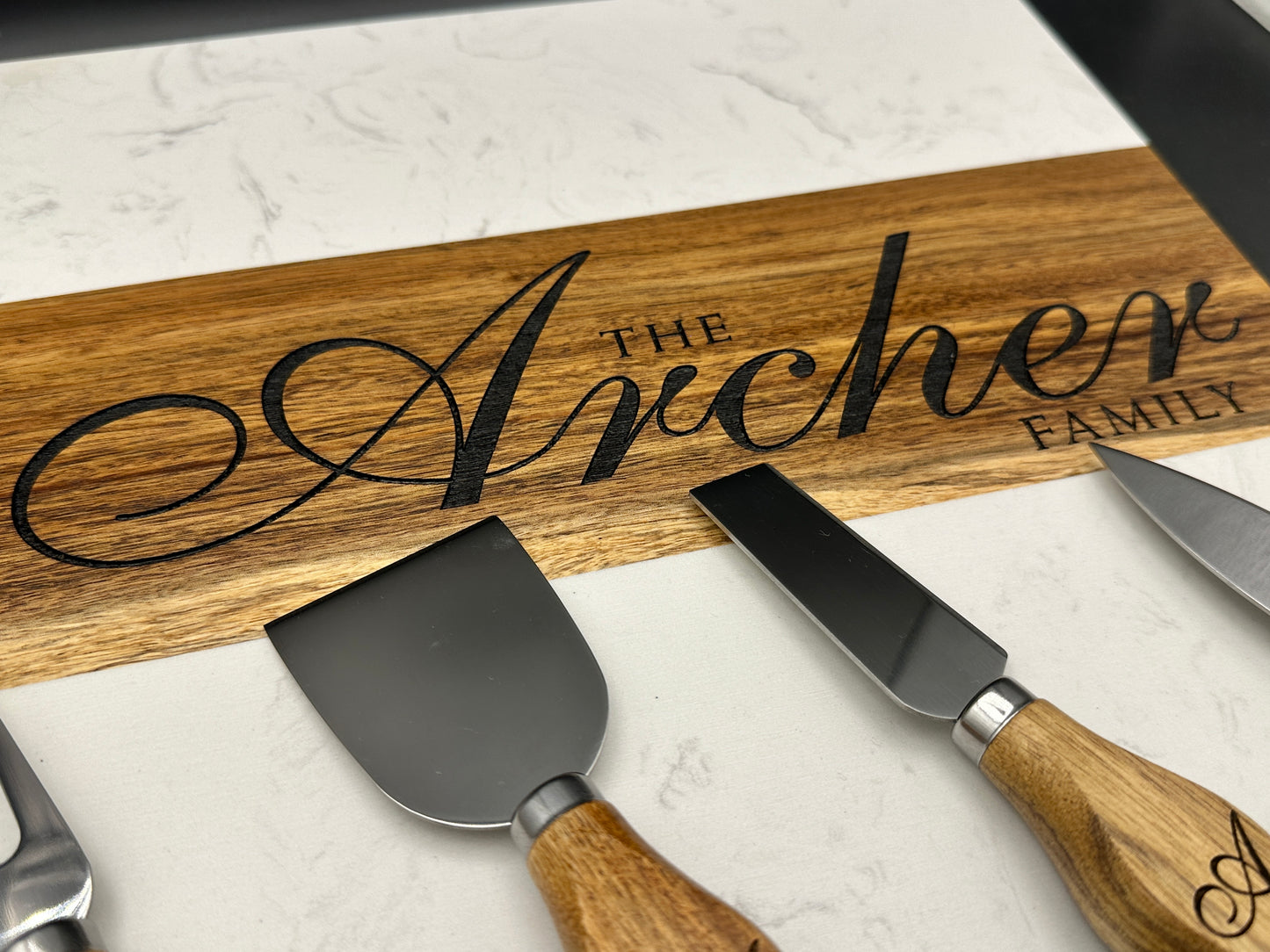 Wooden cutting board with 'The Archer Family' engraving and cheese knives on a marble surface.